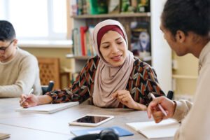 This image shows a focused person in a beige hijab collaborating with classmates in a classroom setting.