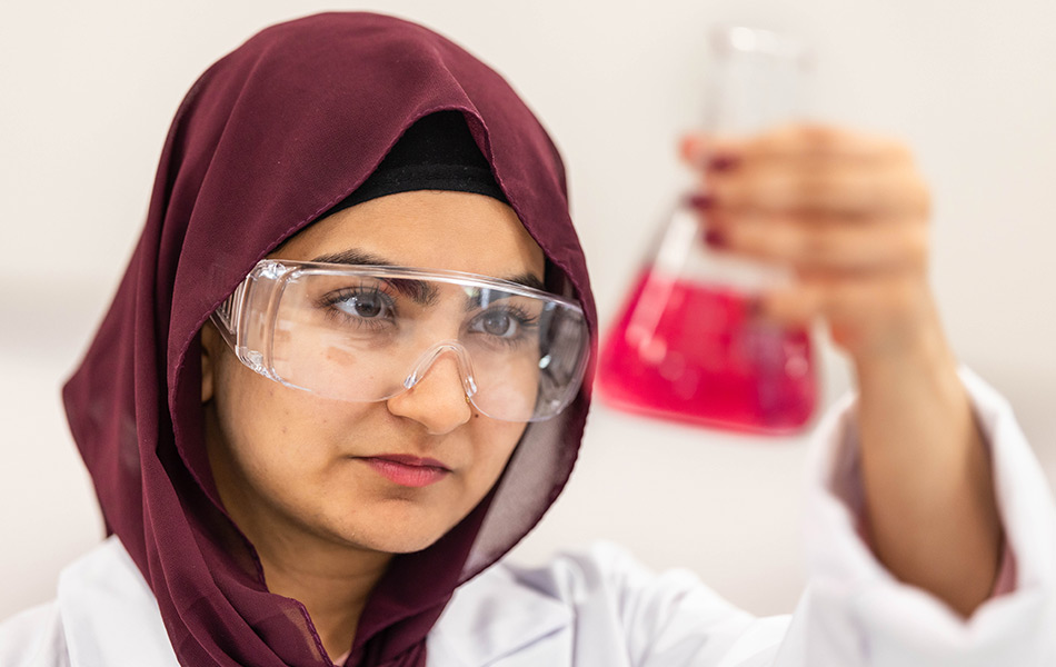 Young girl dressed in white lab coat examining a conical glass with red liquid inside